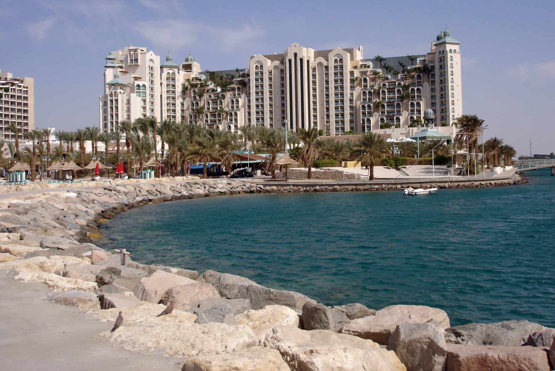 Coastal cityscape featuring modern apartment buildings along a rocky shoreline with palm trees and clear skies.