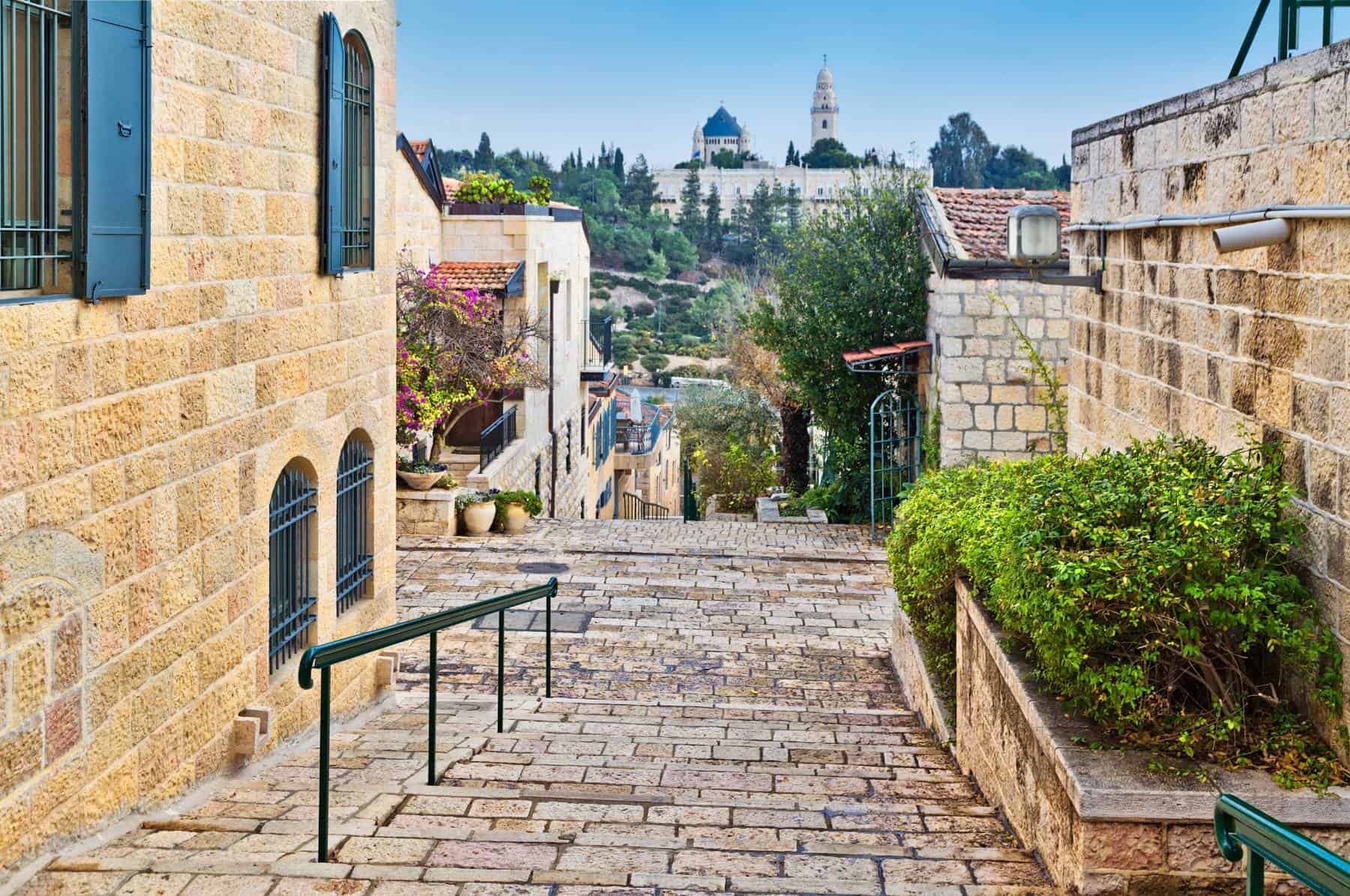 Stone stairway leading down a narrow alley flanked by traditional stone buildings with greenery and a clear blue sky, with a historic domed structure visible in the distance.