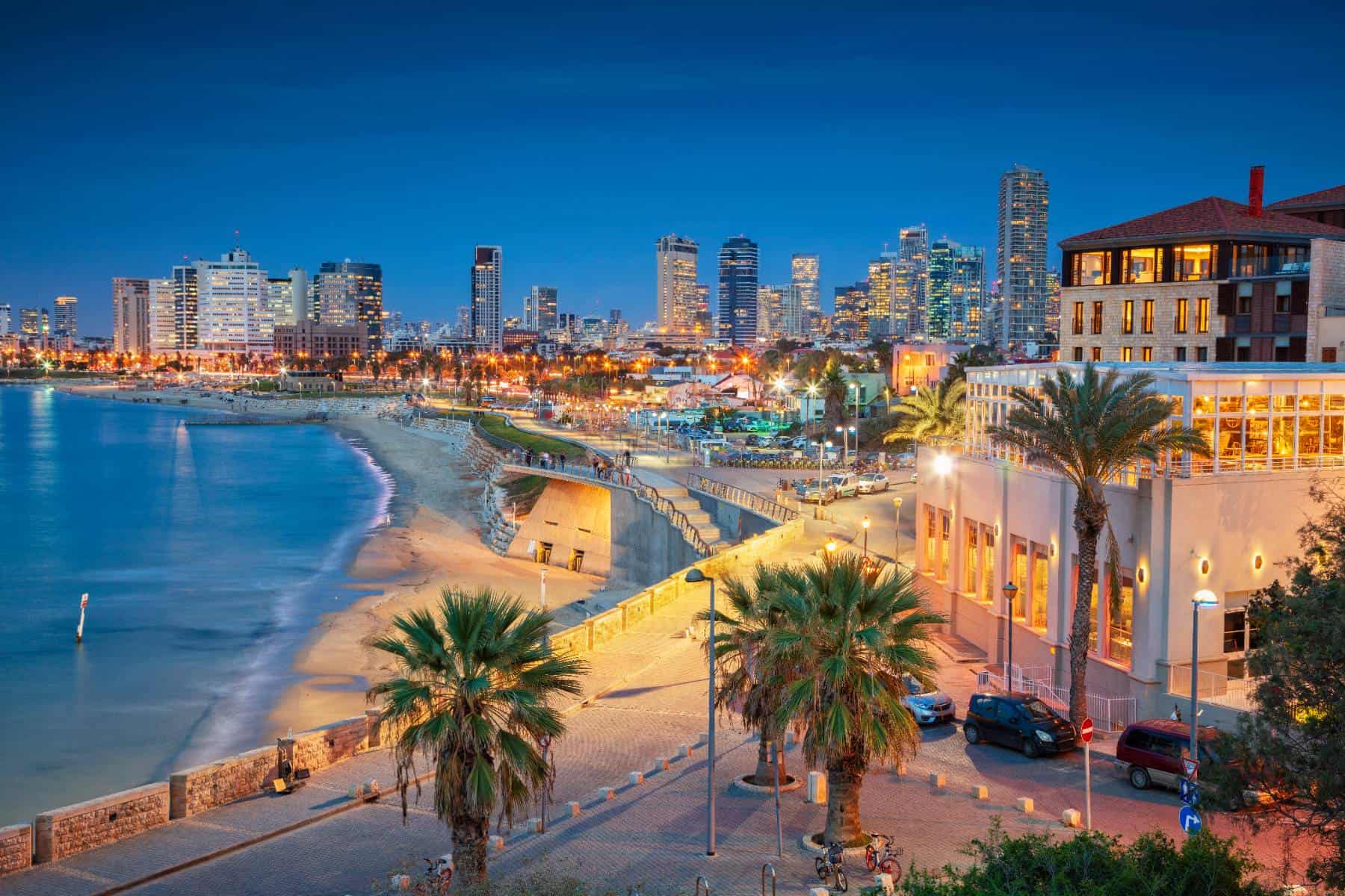 Twilight cityscape overlooking a coastal promenade with illuminated buildings and a serene beachfront.