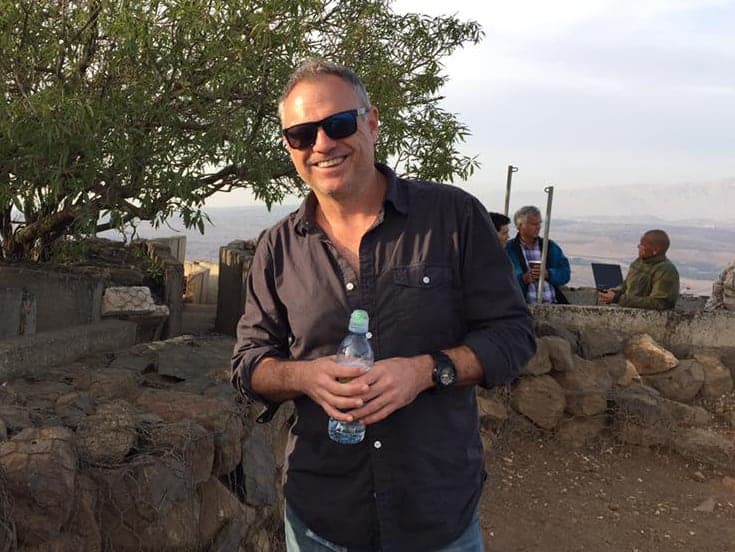 A man wearing sunglasses and a dark shirt holds a water bottle while standing outdoors near a tree and stone wall. There are other people in the background near some equipment.